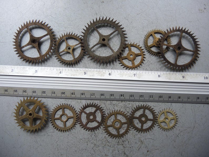 May include: A collection of antique brass gears of varying sizes, arranged on a gray surface. The gears have a weathered, aged appearance, with visible rust and discoloration. A ruler is placed next to the gears for scale, showing measurements in centimetres.