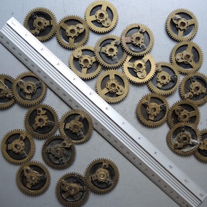 May include: A collection of small, brass-colored clock gears scattered on a surface. A silver ruler is placed diagonally across the gears, providing a size reference. The gears have intricate designs and are likely vintage or antique.