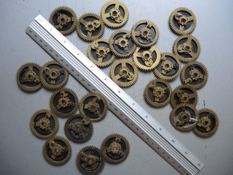 May include: A collection of small, brass-colored clock gears scattered on a surface. A silver ruler is placed diagonally across the gears, providing a size reference. The gears have intricate designs and are likely vintage or antique.
