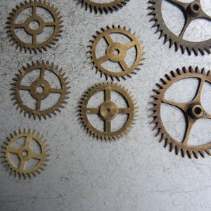 May include: A collection of small, brass-coloured gears with detailed teeth and central holes. The gears are of varying sizes and are arranged on a textured, grey surface, evoking a mechanical or industrial aesthetic.