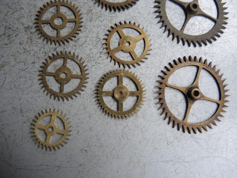 May include: A collection of small, brass-coloured gears with detailed teeth and central holes. The gears are of varying sizes and are arranged on a textured, grey surface, evoking a mechanical or industrial aesthetic.