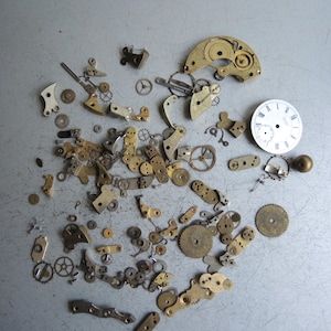 May include: A selection of disassembled watch components, such as cogs, springs, and plates, arranged on a grey surface. The parts are in various sizes and shapes, with brass and silver hues. A white watch face with black numerals is also visible.