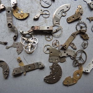 K&ouml;nnte beinhalten: Eine Sammlung antiker Uhrenteile, darunter Zahnr&auml;der, Hebel und Platten, in verschiedenen Formen und Gr&ouml;&szlig;en. Die Komponenten bestehen aus Metall und weisen Alters- und Abnutzungserscheinungen auf, mit einer Mischung aus Silber- und Goldt&ouml;nen.