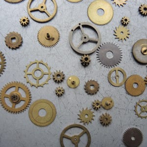 May include: A collection of various sized brass and silver-toned gears scattered on a gray surface. The gears have different spoke and hole designs, with some having a circular or square center.