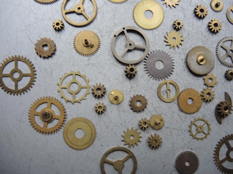 May include: A collection of various sized brass and silver-toned gears scattered on a gray surface. The gears have different spoke and hole designs, with some having a circular or square center.