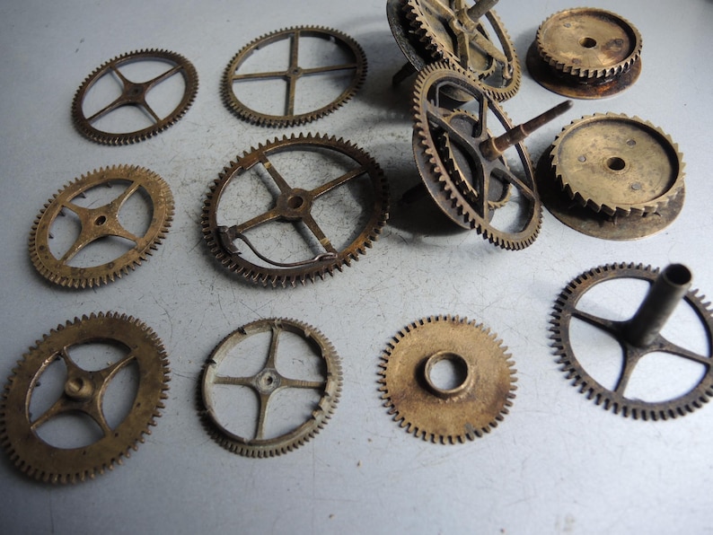 May include: A collection of antique clock gears in various sizes and designs. The gears are made of brass and have a worn, aged appearance. Some gears have spokes, while others are solid discs. The image shows a variety of shapes and sizes.