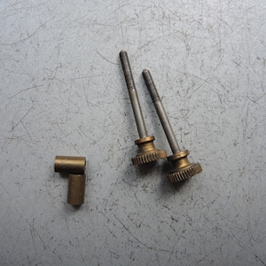 Puede incluir: Dos tornillos metálicos con perillas de color latón y una pequeña pieza cilíndrica de color latón. Los tornillos tienen un vástago roscado y un diseño similar a un engranaje en la perilla. Las piezas están sobre una superficie gris.