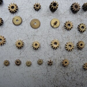 May include: A selection of small brass gears of different sizes, arranged on a grey surface. The gears are circular with teeth around the edge and a central hole. Some gears have a small post protruding from the centre.