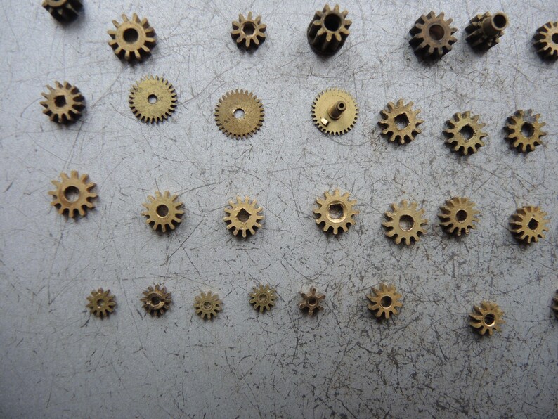May include: A selection of small brass gears of different sizes, arranged on a grey surface. The gears are circular with teeth around the edge and a central hole. Some gears have a small post protruding from the centre.