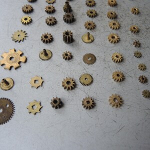 May include: A selection of small, antique brass gears in various sizes and shapes. The gears are arranged on a light grey surface, highlighting their intricate designs and aged patina. Suitable for steampunk or vintage-themed projects.