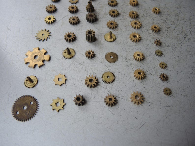 May include: A selection of small, antique brass gears in various sizes and shapes. The gears are arranged on a light grey surface, highlighting their intricate designs and aged patina. Suitable for steampunk or vintage-themed projects.