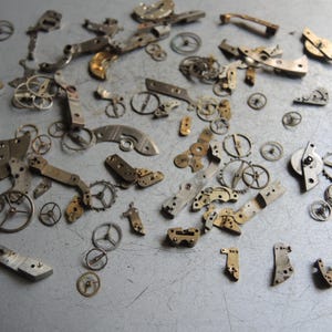 K&ouml;nnte beinhalten: Eine Sammlung antiker Uhrenteile, darunter Zahnr&auml;der, Hebel und Platten, auf einer metallischen Oberfl&auml;che verstreut. Die Komponenten sind haupts&auml;chlich silber- und goldfarben und zeigen komplizierte mechanische Details. Ideal f&uuml;r Steampunk oder Schmuckherstellung.