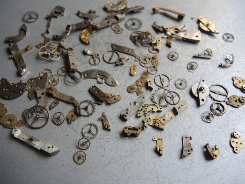 K&ouml;nnte beinhalten: Eine Sammlung antiker Uhrenteile, darunter Zahnr&auml;der, Hebel und Platten, auf einer metallischen Oberfl&auml;che verstreut. Die Komponenten sind haupts&auml;chlich silber- und goldfarben und zeigen komplizierte mechanische Details. Ideal f&uuml;r Steampunk oder Schmuckherstellung.