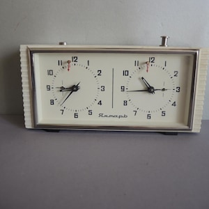 May include: A rectangular analog chess clock with a cream-colored casing and two circular clock faces. Each face features black numerals from 1 to 12, black hands, and a small red indicator. The word "Янтарь" is printed in black on the front.