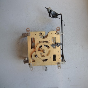 May include: A vintage clock mechanism with brass gears and a decorative black bird perched on a metal rod. The clock mechanism features intricate gears and a rectangular brass plate.