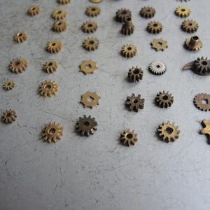 May include: A selection of small, brass-coloured gears of varying sizes and designs scattered on a grey surface. The gears feature different numbers of teeth and shapes, suitable for watchmaking or mechanical projects.