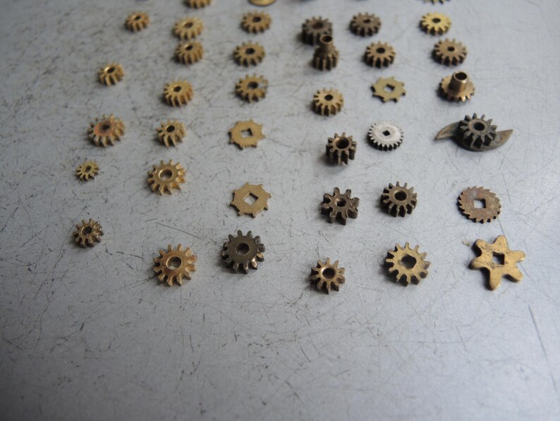 May include: A selection of small, brass-coloured gears of varying sizes and designs scattered on a grey surface. The gears feature different numbers of teeth and shapes, suitable for watchmaking or mechanical projects.