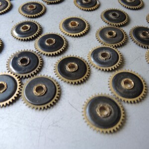 May include: A collection of small, circular gears with a brass-colored outer ring and a dark center. The gears have a toothed edge and a central hole. The gears are scattered across a light-colored surface.