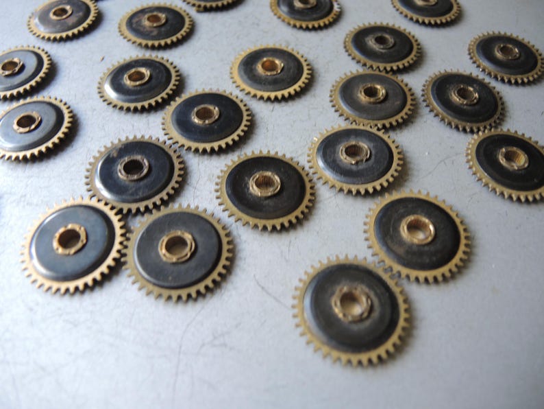 May include: A collection of small, circular gears with a brass-colored outer ring and a dark center. The gears have a toothed edge and a central hole. The gears are scattered across a light-colored surface.