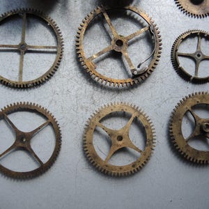 May include: A collection of antique brass clock gears of various sizes. The gears have intricate teeth and spoke designs, showcasing their mechanical complexity. The gears are arranged on a gray surface, highlighting their aged appearance.