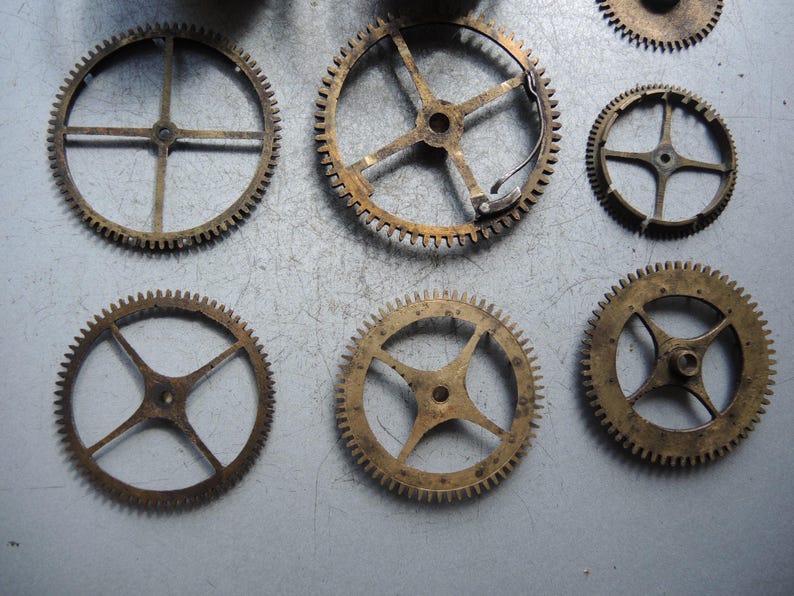 May include: A collection of antique brass clock gears of various sizes. The gears have intricate teeth and spoke designs, showcasing their mechanical complexity. The gears are arranged on a gray surface, highlighting their aged appearance.