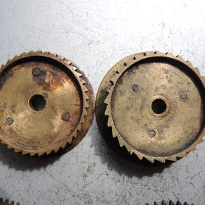 May include: Two antique brass gears with a central hole and toothed edges. The gears show signs of age and wear, with some areas of discoloration and rust. The gears are likely from a clock or other mechanical device.