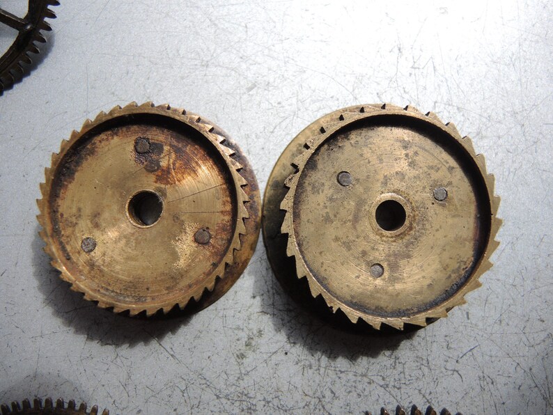 May include: Two antique brass gears with a central hole and toothed edges. The gears show signs of age and wear, with some areas of discoloration and rust. The gears are likely from a clock or other mechanical device.