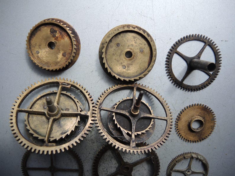 May include: A collection of antique brass clock gears of various sizes and designs. The gears have a worn, aged appearance with visible teeth and central holes. Some gears have spokes, while others are solid.
