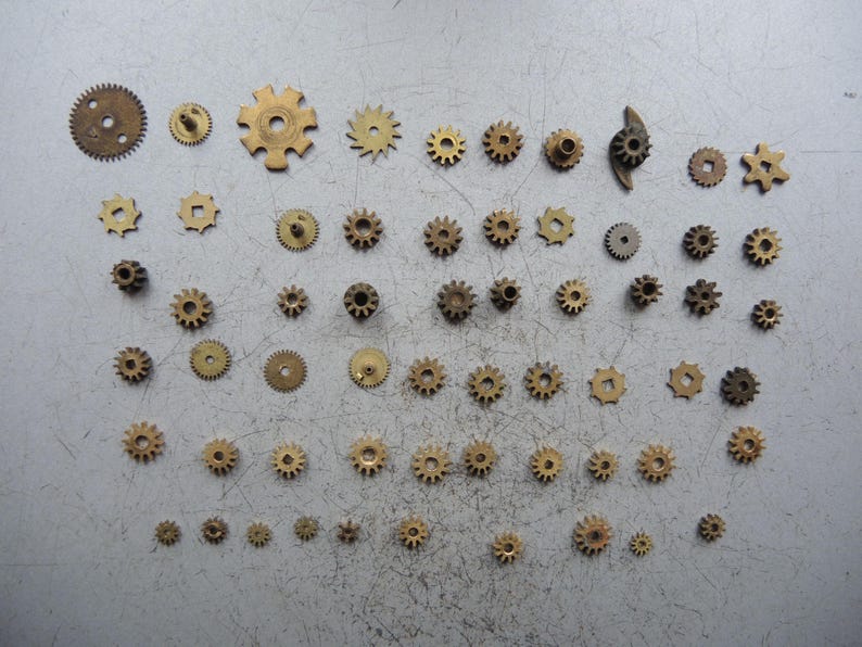 May include: A selection of small brass gears, each with a unique design and size, arranged on a light grey surface. The gears display signs of age, with some showing intricate teeth patterns. Measurements vary, with the largest gear approximately 2.5 cm in diameter.