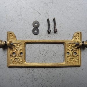 Könnte beinhalten: Ein antiker Schubladengriff aus Messing mit aufwendigen floralen Details, zwei Schrauben und zwei Unterlegscheiben. Der rechteckige Griff hat eine zentrale Öffnung und dekorative Knöpfe an jedem Ende. Die Hardware ist wahrscheinlich für einen Schrank oder eine Schublade.