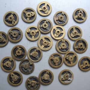 May include: A collection of small, brass-colored clock gears with intricate designs. The gears have a circular shape with teeth around the edges and various internal mechanisms. They are scattered on a light gray surface.