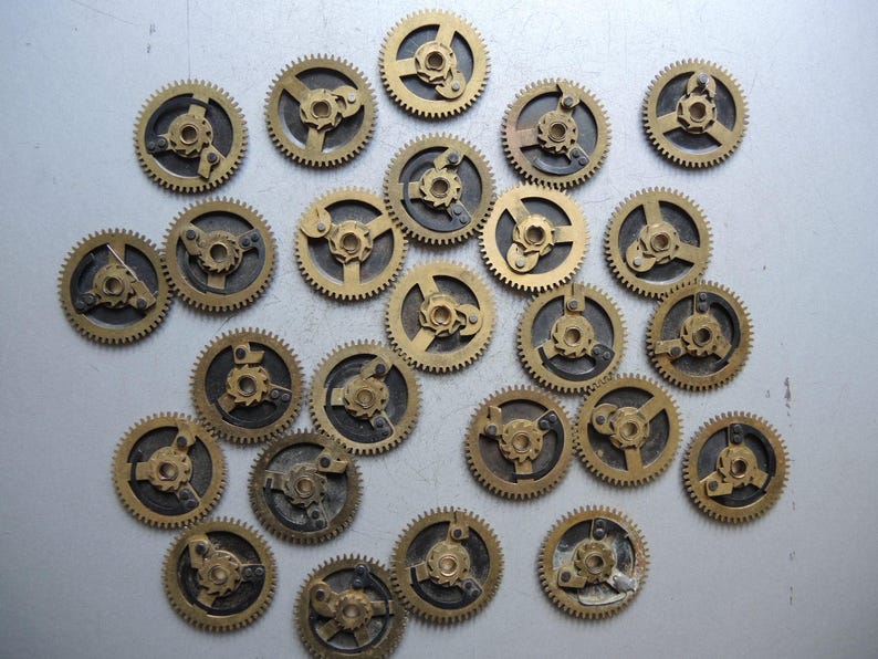 May include: A collection of small, brass-colored clock gears with intricate designs. The gears have a circular shape with teeth around the edges and various internal mechanisms. They are scattered on a light gray surface.