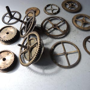 May include: A collection of antique brass clock gears of various sizes and designs. The gears have intricate teeth and spoke patterns, showcasing a vintage aesthetic. Some gears have cylindrical posts, while others are flat. The gears are arranged on a light-colored surface.