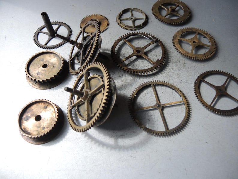 May include: A collection of antique brass clock gears of various sizes and designs. The gears have intricate teeth and spoke patterns, showcasing a vintage aesthetic. Some gears have cylindrical posts, while others are flat. The gears are arranged on a light-colored surface.