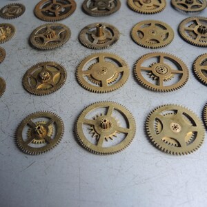 May include: A collection of small, brass-colored clock gears of various sizes. The gears have intricate teeth and central hubs, arranged on a light gray surface. The gears are likely vintage or antique watch parts.
