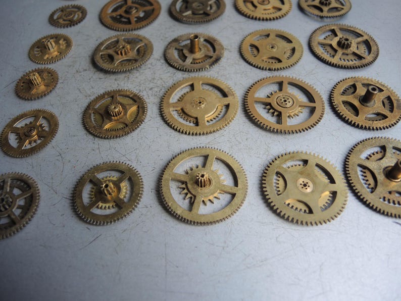 May include: A collection of small, brass-colored clock gears of various sizes. The gears have intricate teeth and central hubs, arranged on a light gray surface. The gears are likely vintage or antique watch parts.