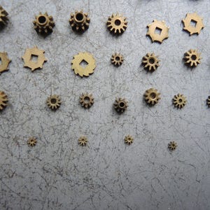 May include: A collection of small, brass-colored gears of varying sizes. The gears have different tooth designs and center hole shapes, scattered on a gray surface. The gears are likely for a mechanical or clockwork project.