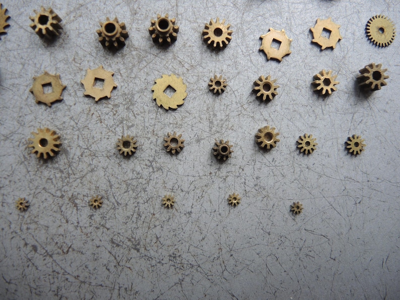 May include: A collection of small, brass-colored gears of varying sizes. The gears have different tooth designs and center hole shapes, scattered on a gray surface. The gears are likely for a mechanical or clockwork project.