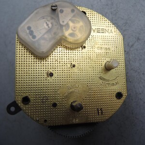 Op de afbeelding: Een vintage uurwerk met een goudkleurige, geperforeerde metalen basis. Een doorzichtige plastic hoes omsluit tandwielen en andere interne componenten. Het woord "VESNA" is zichtbaar, samen met andere markeringen en een klein tandwiel.