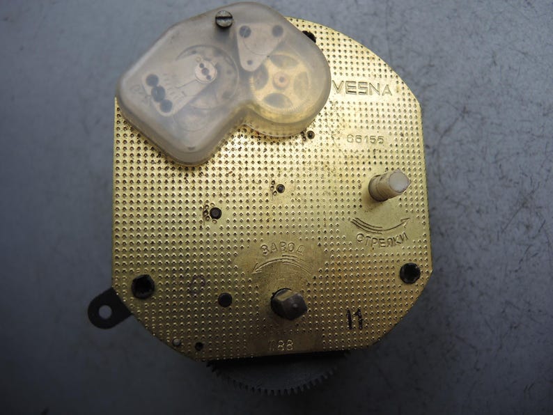 Op de afbeelding: Een vintage uurwerk met een goudkleurige, geperforeerde metalen basis. Een doorzichtige plastic hoes omsluit tandwielen en andere interne componenten. Het woord "VESNA" is zichtbaar, samen met andere markeringen en een klein tandwiel.