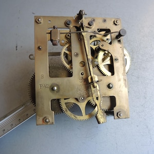 Può includere: Un movimento di orologio vintage in ottone con ingranaggi e meccanismo a pendolo visibili. Il telaio rettangolare mostra il testo "Silesia" e "P42". Un righello è posizionato accanto all'orologio.