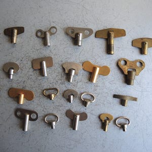 Pode incluir: Uma coleção de chaves de relógio vintage em várias formas, incluindo designs em T, circulares e retangulares. As chaves são feitas de latão e metal prateado, dispostas sobre uma superfície cinza.