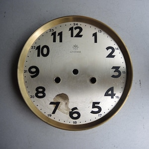 Puede incluir: Una esfera de reloj vintage con un borde dorado y un centro plateado. Los números negros marcan las horas, con la marca "JUNGHANS" visible. La esfera muestra signos de desgaste, incluyendo una zona dañada cerca de los números 6 y 7.