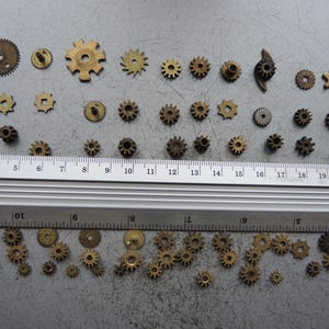 May include: A selection of small, brass-toned gears in diverse shapes and sizes, laid out on a grey surface. A ruler with centimetre markings is positioned across the gears, offering scale. These gears are likely components for a clock or mechanical device.