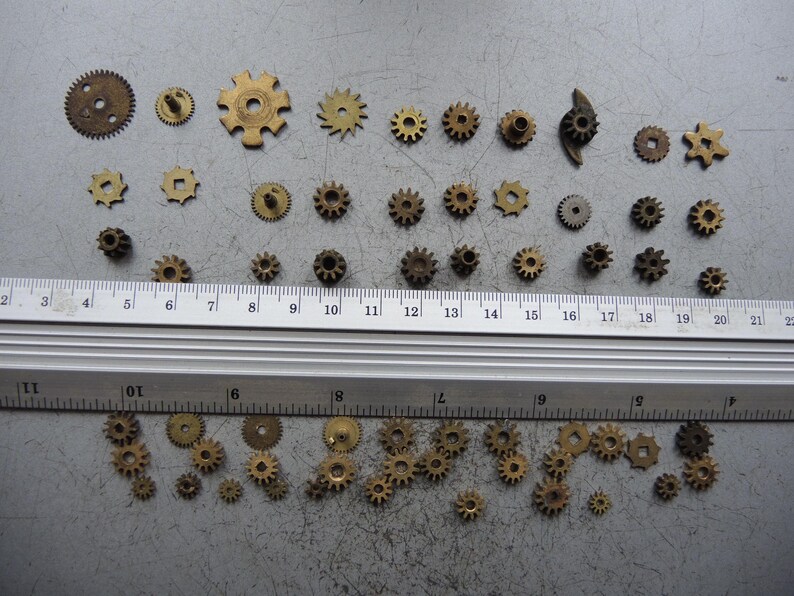 May include: A selection of small, brass-toned gears in diverse shapes and sizes, laid out on a grey surface. A ruler with centimetre markings is positioned across the gears, offering scale. These gears are likely components for a clock or mechanical device.