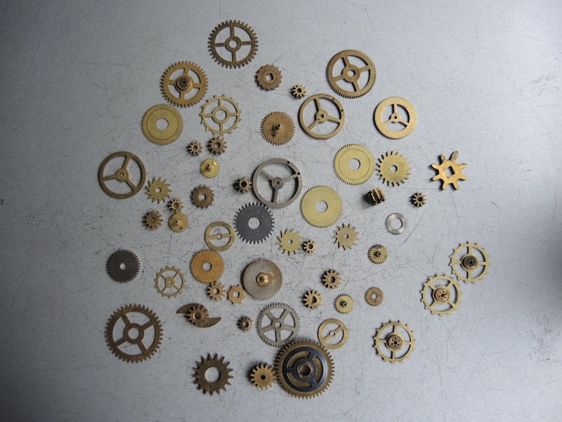 May include: A collection of various sized brass and steel gears scattered on a gray surface. The gears have different shapes and tooth counts, suggesting a mechanical or steampunk theme.