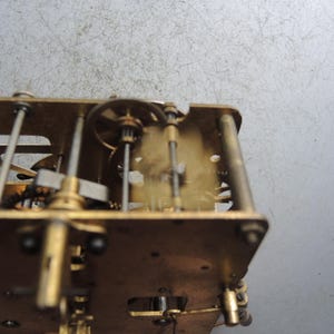 May include: Close-up of a clock mechanism with brass gears, rods, and plates. The intricate internal workings of the clock are visible, showcasing the mechanical components and their arrangement. The clock's frame is rectangular, with various gears and levers.