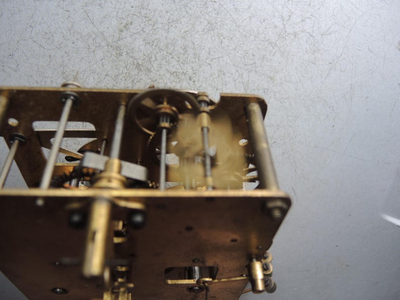 May include: Close-up of a clock mechanism with brass gears, rods, and plates. The intricate internal workings of the clock are visible, showcasing the mechanical components and their arrangement. The clock's frame is rectangular, with various gears and levers.