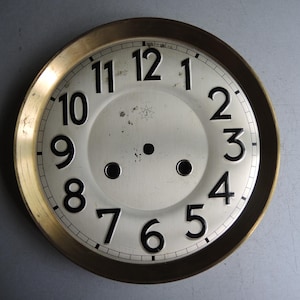 Può includere: Un quadrante di orologio vintage con un bordo color oro e un centro color crema. I numeri neri segnano le ore, con due piccoli fori e un disegno a forma di stella al centro. Il quadrante dell'orologio è di forma circolare.