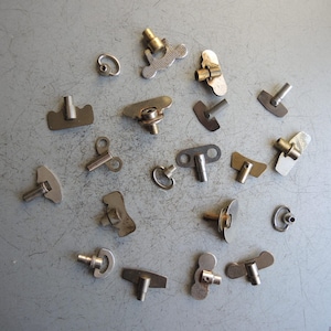 Op de afbeelding: Een verzameling vintage metalen kloksleutels en opwindmechanismen, in verschillende vormen en maten. De stukken zijn in tinten zilver, goud en brons, verspreid over een grijze ondergrond. Sommige hebben textuurpatronen.